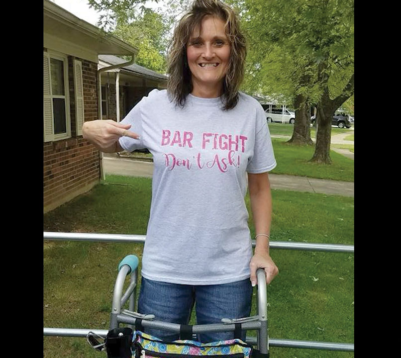 Beth Waddell stands with her walker in front of her house after her car accident wearing a 'bar fight don't ask' shirt