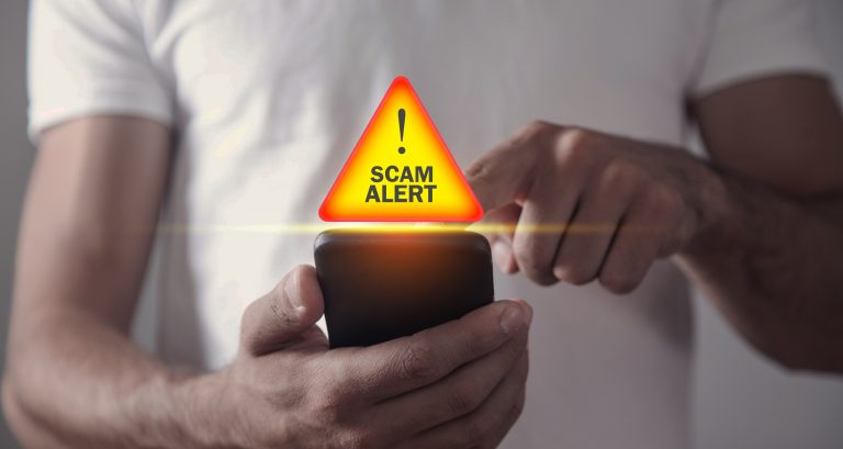 White man holding a cell phone with an illustration of a scam alert badge. Many online scams offer users a fake cash reward.