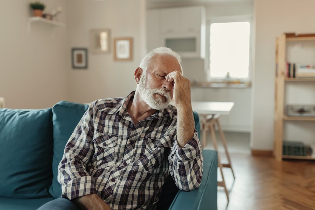 White man with a gray beard sitting on a couch, pinching the bridge of his nose and appearing in pain. Car accident injury symptoms are sometimes subtle.