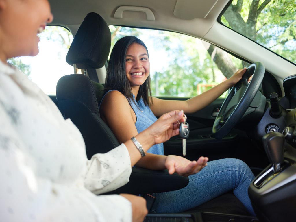 A side view photo of a teenage driver in a car as her mother sits next to her and hands her the keys to drive.